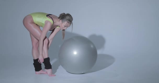 Beautiful girl in a yellow sports suit and black leggings is engaged in gymnastics on a gray rubber ball, swimsuit, beautiful skin, figure, barefoot, long hair in the tail - Video