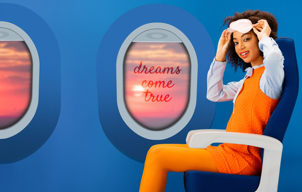 smiling african american woman in orange retro dress holding sleeping mask and sitting on seat on blue background with portholes and dreams come true illustration - Photo, Image