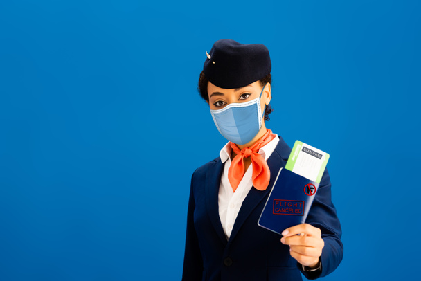 auxiliar de vuelo afroamericano en máscara médica con pasaporte y billete de avión aislado en azul
  - Foto, imagen