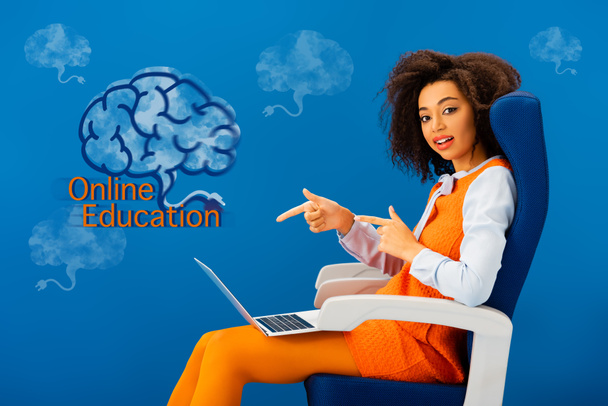 donna afroamericana seduta sul sedile e che punta con le dita al computer portatile su sfondo blu con illustrazione educazione online
 - Foto, immagini