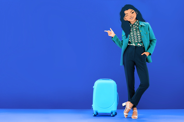 Mavi arka planda seyahat çantasının yanında duran resimli yüzü olan kadın parmağıyla işaret ediyor. - Fotoğraf, Görsel