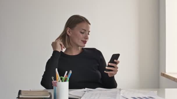 An attractive young blonde businesswoman is looking at her smartphone at a working time indoors in a white office - Кадри, відео