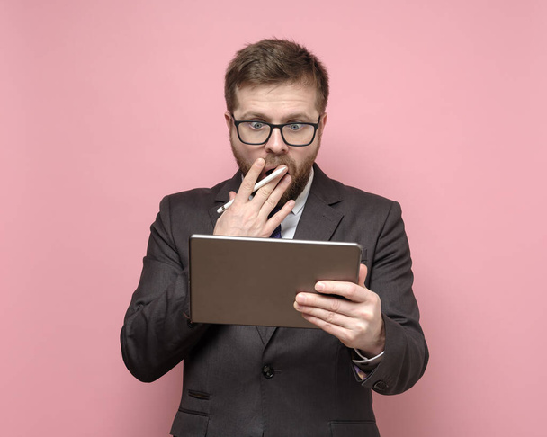 Excited man in a suit looks at the tablet in surprise, covering mouth with hand. - Foto, imagen