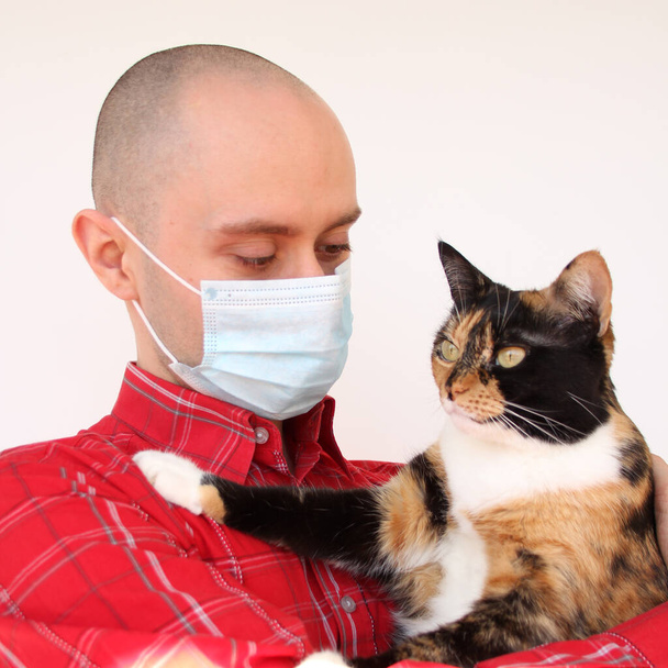 young man in a white medical antivirus mask holds a domestic dark cat in his hands, the concept of coronavirus COVID-19 in humans and animals, veterinary medicine - Foto, imagen