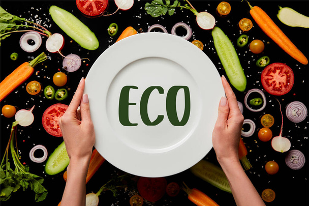 bijgesneden weergave van vrouw met ronde plaat met eco illustratie op plantaardige patroon achtergrond geïsoleerd op zwart - Foto, afbeelding