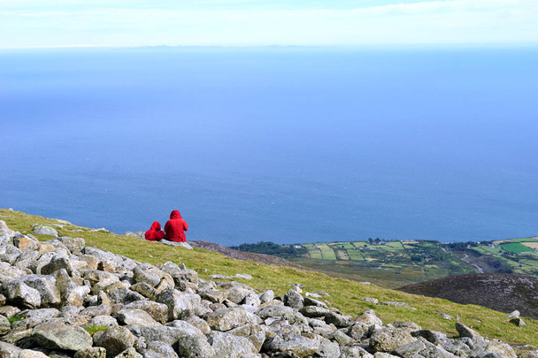 Δύο τουρίστες με κόκκινα παλτά στην κορυφή του βουνού Slieve Donard στα βουνά Mourne, Βόρεια Ιρλανδία - Φωτογραφία, εικόνα