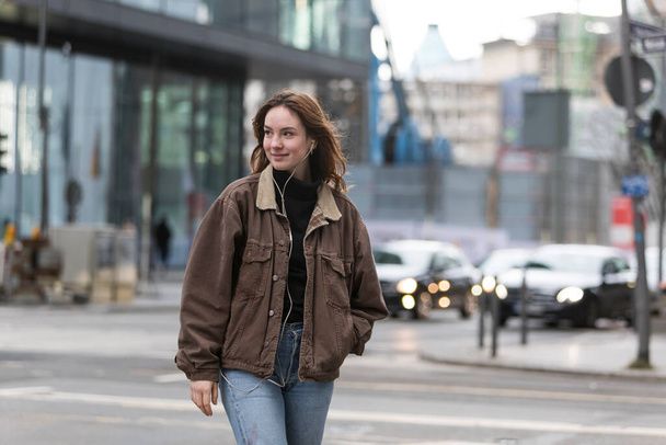 Молода жінка слухає навушники, посміхаючись головою, що йде по міській вулиці
. - Фото, зображення
