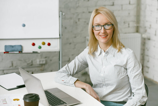 Smiling businesswoman works laptop in office. Charming blonde woman smiles looking at camera while sitting at office desk with computer on it - 写真・画像