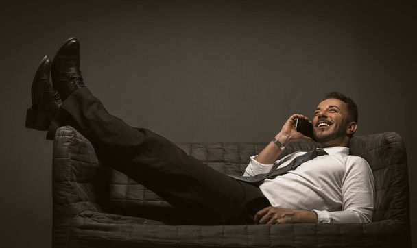 Businessman with mobile lies throwing legs on the sofa. Smiling man lying at couch cross-legged talking over smartphone on grey back. Business communication concept - Valokuva, kuva