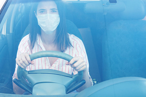 Młoda kobieta w ochronnej masce medycznej kierująca samochodem patrzy w kamerę. Osoba w masce. Bezpieczeństwo podczas pandemii koronawirusa, epidemii covid-19.  - Zdjęcie, obraz