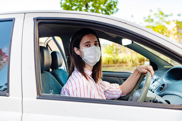 Junge Frau in medizinischer Schutzmaske am Steuer eines Autos blickt in die Kamera. Person mit Maske. Sicherheit während der Coronavirus-Pandemie, Epidemie covid-19.  - Foto, Bild