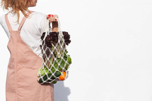 軽い夏服の若い女性は、ビニール袋のない野菜、緑とメッシュショッピングバッグを保持しています。廃棄物ゼロ、プラスチックフリー環境に優しいコンセプト。手作りのマクラームバッグ。持続可能なライフスタイル.   - 写真・画像