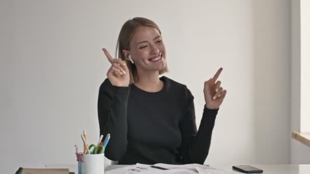 A happy young blonde businesswoman is listening to music and dancing while having success at a working time indoors in a white office - Video, Çekim