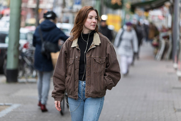 Young caucasian woman in jeans and coat listening to earphones walking on sidewalk. Looking away. - Photo, Image
