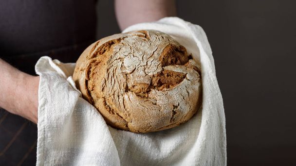 Hands with fresh rye-wheat bread on a cotton towel. Dark background wirh copy space - Foto, Bild