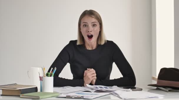 Una giovane donna bionda sorpresa si sta rallegrando e sta facendo un gesto vincente seduta a tavola al chiuso in un ufficio bianco
 - Filmati, video