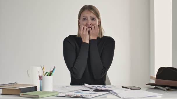 Eine schockierte, verängstigte junge blonde Frau macht sich Sorgen, während sie in einem weißen Büro am Tisch sitzt - Filmmaterial, Video