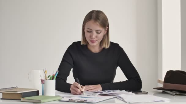 Μια όμορφη νεαρή ξανθιά γυναίκα γράφει κάτι, ενώ έρχεται με μια καλή ιδέα κάθεται στο τραπέζι μέσα σε ένα λευκό γραφείο - Πλάνα, βίντεο
