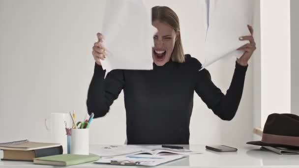 Eine wütende junge blonde Frau schreit und wirft Papierdokumente hinaus, während sie drinnen in einem weißen Büro am Tisch sitzt - Filmmaterial, Video