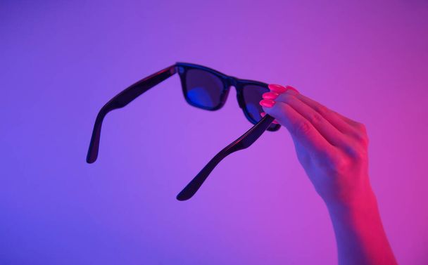 Black sunglasses in a female hand with bright orange manicure in a trendy neon light. - Foto, Bild