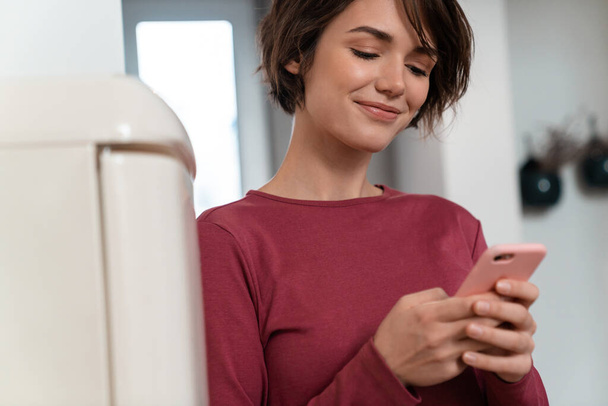 Photo of joyful young woman in leisure clothes smiling and using smartphone while leaning on fridge at kitchen - Fotoğraf, Görsel