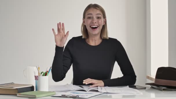 Szczęśliwa pozytywna młoda blondynka macha ręką robiąc gest powitania siedząc przy stole w białym biurze - Materiał filmowy, wideo