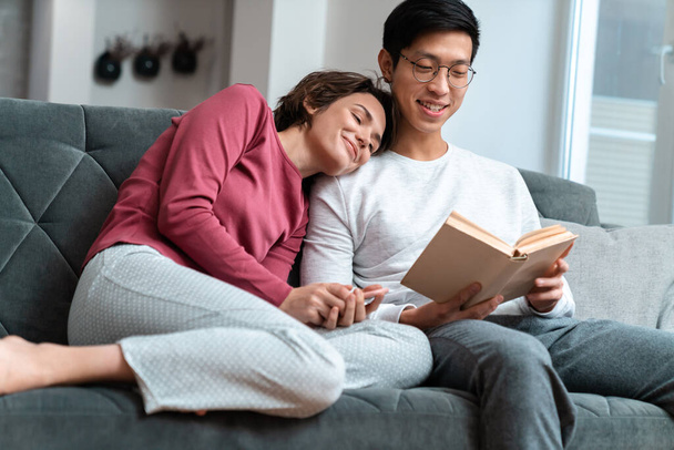 Photo of pleased multinational couple reading book and using smartphone while sitting on sofa at home - Фото, изображение