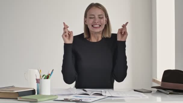 Eine aufgeregte junge blonde Frau drückt die Daumen, während sie in einem weißen Büro am Tisch sitzt und einen Wunsch äußert - Filmmaterial, Video