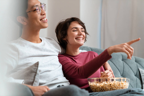 Photo of joyful multinational couple eating popcorn and watching movie while sitting on sofa at home - Fotó, kép