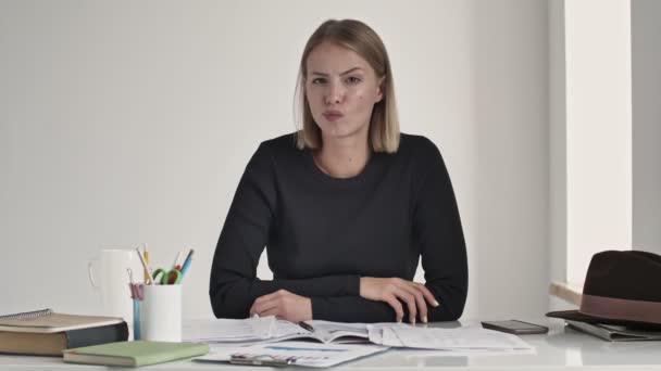 A serious young blonde woman is shaking her head negative while disagreeing with something sitting at the table indoors in a white office - Video, Çekim