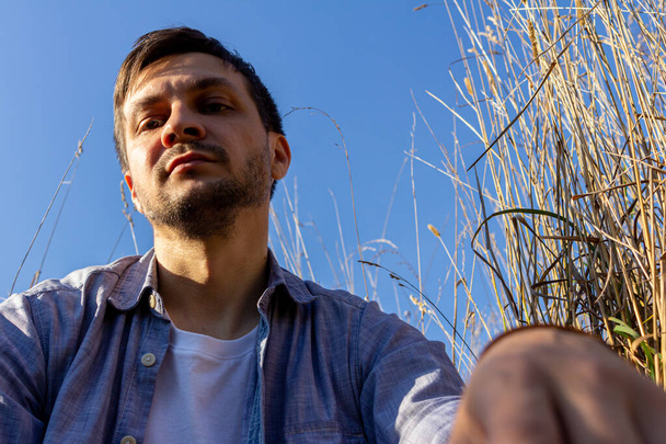 a young attractive man wearing jeans,  white T-shirt and a blue shirt lies on a field of cereals against a blue sky and looks to camera - Foto, Imagen
