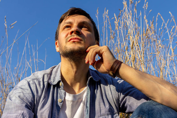 Ένας νεαρός ελκυστικός άντρας που φοράει τζιν, ένα λευκό μπλουζάκι και ένα μπλε πουκάμισο κείτεται σε ένα χωράφι δημητριακών και κοιτάζει ψηλά.. - Φωτογραφία, εικόνα