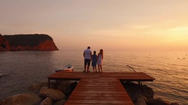 Egy család áll egy fa tengeri mólón, kéz a kézben, csodálva a színes tüzes naplementét a tenger felett. A családi értékek, a szeretet és az egység fogalma. Családi nyaralás az Adrián Montenegróban. - Felvétel, videó