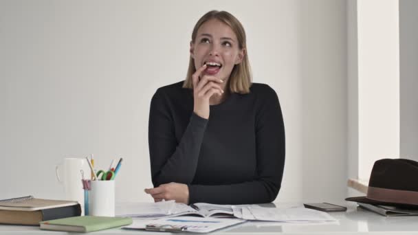 Eine lächelnde junge blonde Frau träumt von etwas, das drinnen in einem weißen Büro am Tisch sitzt - Filmmaterial, Video