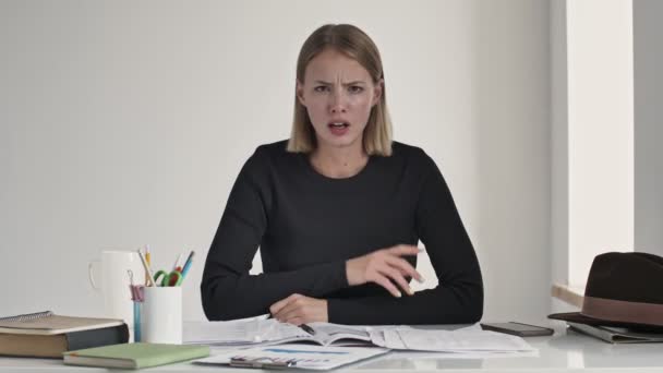 A serious young blonde woman showing a silence gesture sitting at the table indoors in a white office - Filmati, video