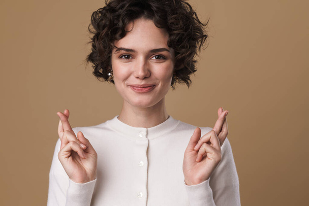 Image of pleased beautiful woman holding fingers crossed foe good luck isolated over beige background - Foto, Bild