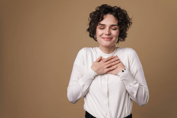 Image of excited beautiful woman smiling and holding hands on her chest isolated over beige background - Fotoğraf, Görsel