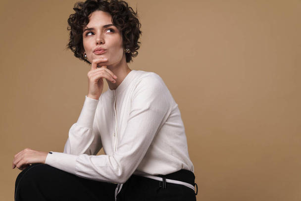 Image of thinking beautiful woman looking upward while sitting isolated over beige background - Fotografie, Obrázek