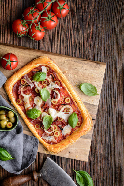 Domowej roboty pizzę z plasterkami bekonu, pomidory, zielone oliwki, ser i cebula, zwieńczone liśćmi bazylii na rustykalnym drewnianym tle - Zdjęcie, obraz