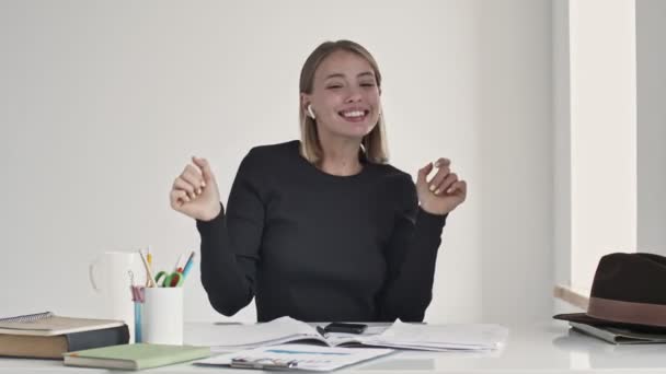 Una joven rubia feliz está disfrutando de la música en sus auriculares sentados en la mesa dentro de una oficina blanca
 - Metraje, vídeo