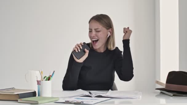 Μια χαρούμενη νεαρή ξανθιά γυναίκα τραγουδάει ένα τραγούδι ακούγοντας τη μουσική στα ακουστικά της να κάθεται στο τραπέζι σε ένα λευκό γραφείο. - Πλάνα, βίντεο