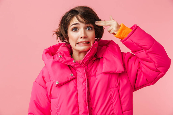 Portrait of young amusing woman in warm coat making gun gesture and looking at camera isolated over pink background - Photo, image