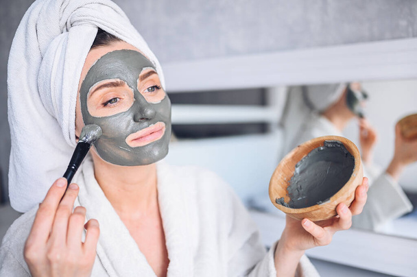 Cosmetology, skin care, face treatment, spa, natural beauty concept. Beautiful smiling woman at home in bathrobe with a towel applying face clay mask against acne to rejuvenate problem skin - Foto, Imagen