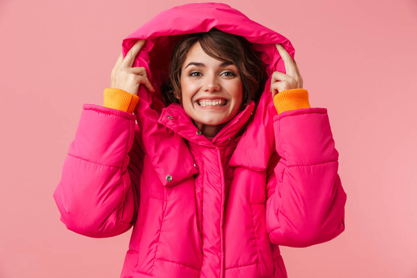 Portrait of young joyful woman in warm hood smiling and looking at camera isolated over pink background - Photo, image