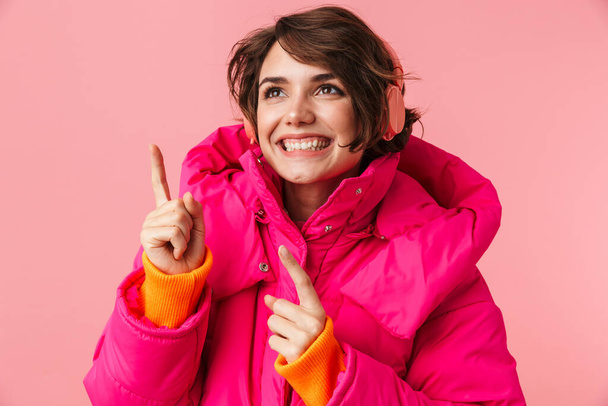 Portrait of young smiling woman in warm coat using headphones and pointing fingers upward isolated over pink background - Foto, Imagem