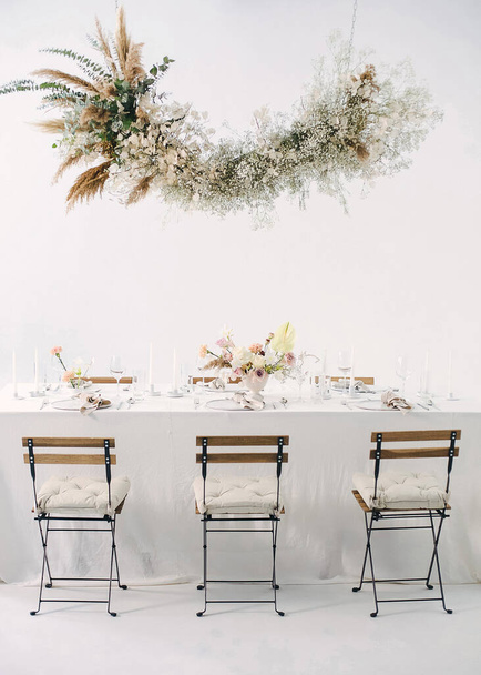 праздничный стол с цветочным букетом, свечами, стульями и цветочной гирляндой
 - Фото, изображение
