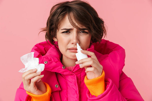 Portrait of unhappy young woman in warm coat holding napkin and using nasal spray isolated over pink background - Fotó, kép