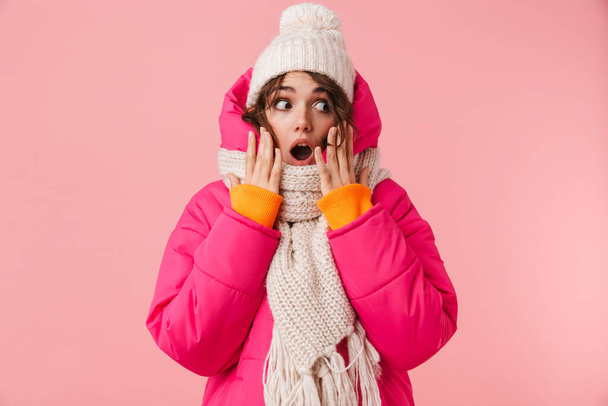 Portrait of beautiful woman in warm clothes expressing surprise and looking aside isolated over pink background - Photo, Image