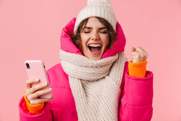 Portrét vzrušené mladé ženy v teplém oblečení křičí a pomocí mobilního telefonu izolované přes růžové pozadí - Fotografie, Obrázek
