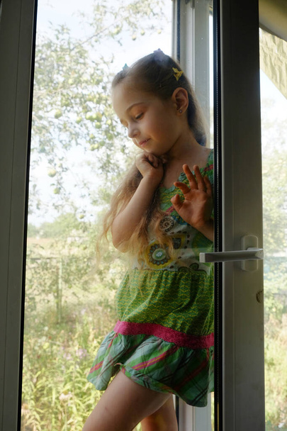 Klein meisje bij het raam. Een 5-jarig meisje zit thuis bij het raam. Jeugd, kind, raam. Blijf thuis. - Foto, afbeelding